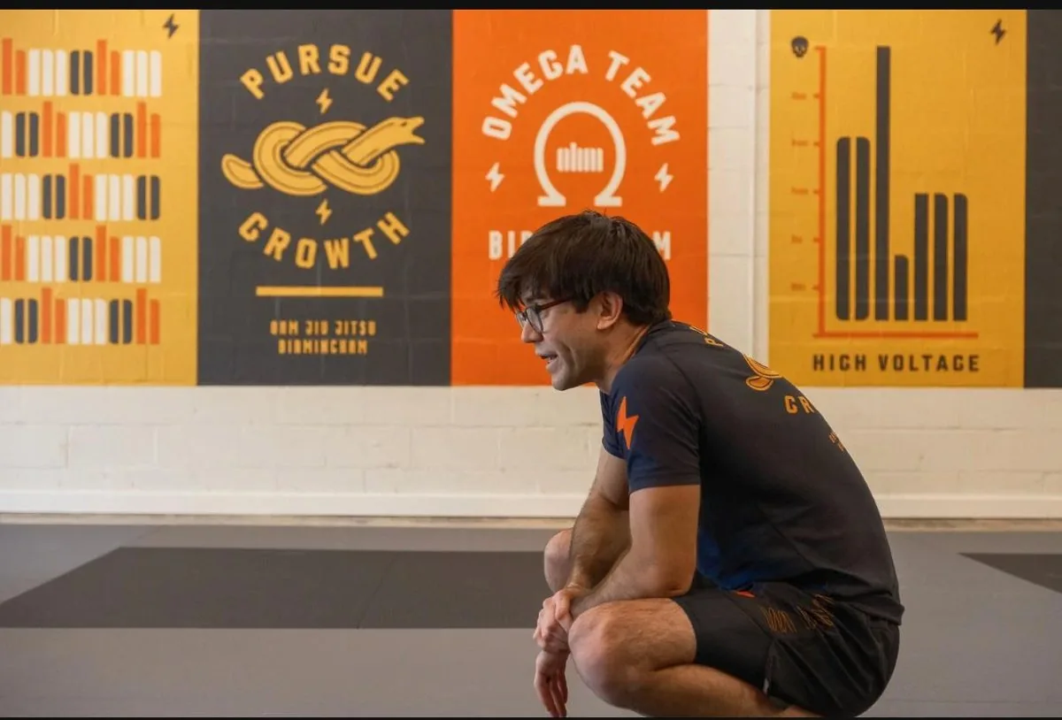 DJ Wright, 2nd degree black belt instructor at Ohm Jiu Jitsu, crouching on the gym floor with motivational posters behind him.