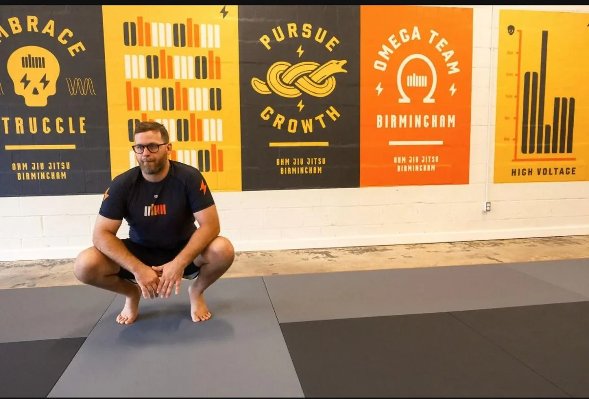 Russell Marbut, 1st degree black belt instructor at Ohm Jiu Jitsu, crouching on a mat with Embrace the Struggle and Pursue Growth posters behind him.